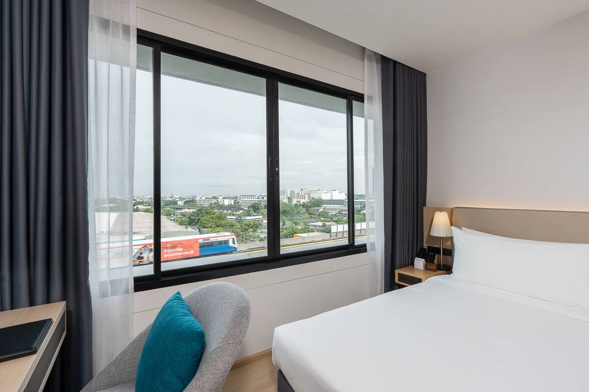 A hotel room with a large window, a bed, a chair, a bedside table, and a nightstand with a lamp.