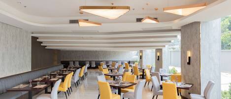 A spacious restaurant with high ceilings, modern pendant lighting, and a mix of yellow and white chairs.