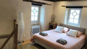 Une chambre à coucher avec un lit, deux fenêtres, un radiateur, une lampe et une poutre en bois au plafond.