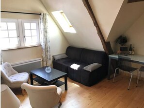 A cozy loft-style room with a black sofa, a small table, and a white armchair. There is a window with curtains, a desk with a computer and a plant, and a wooden ceiling.