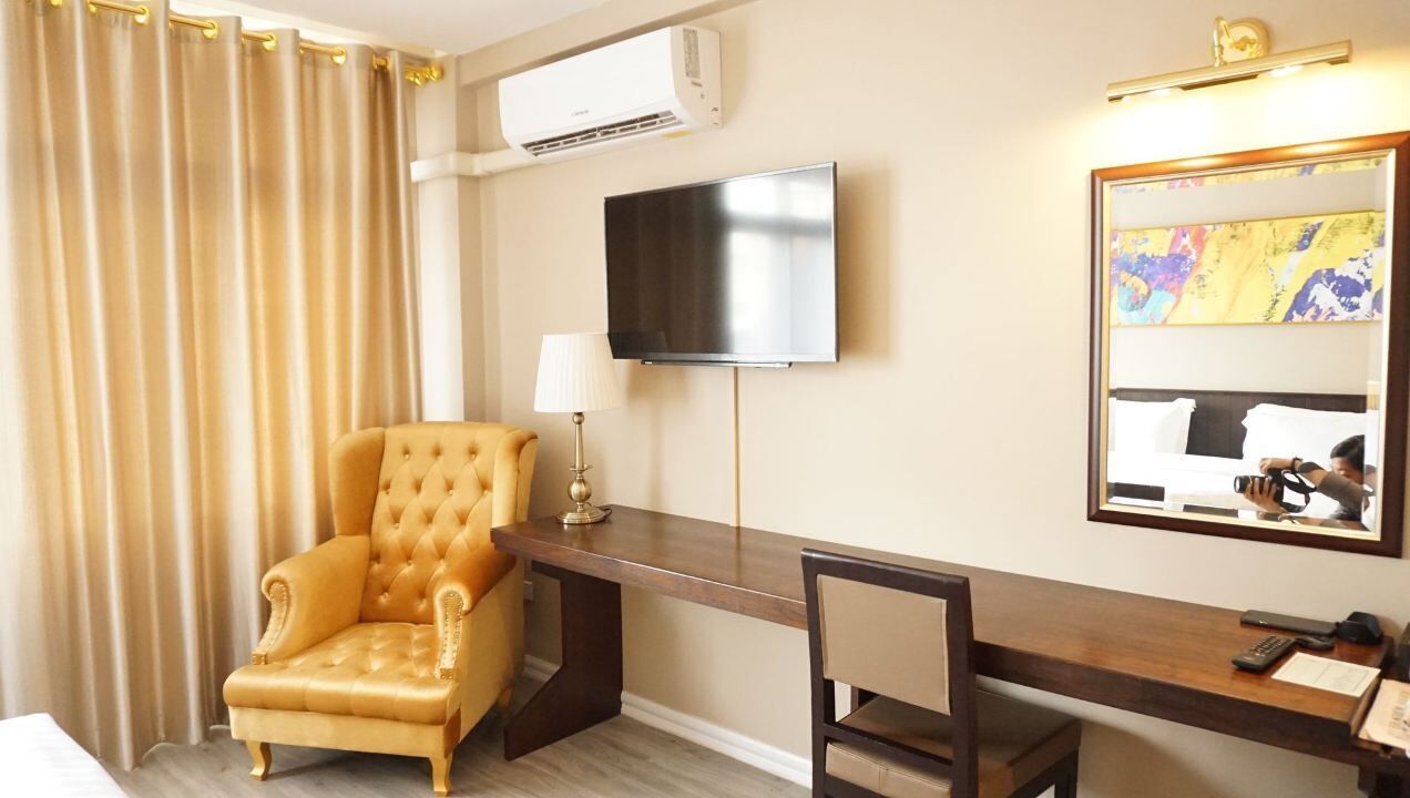 Habitación de hotel con un sillón capitonado, un escritorio con lámpara, un televisor de pantalla plana y un espejo grande que refleja la cama.