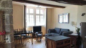 A cozy living room with a wooden coffee table, a grey sofa, and a TV. There's a stone pillar and a window with curtains. - Appartement de Charme "Le Bureau des Armateurs" (Tréguier)