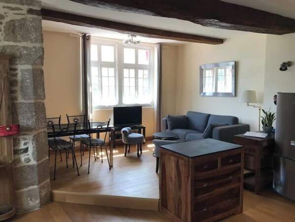 A cozy living room with a wooden coffee table, a grey sofa, and a TV. There's a stone pillar and a window with curtains.