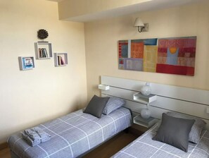 A bedroom with two single beds, a bedside table with a lamp, a wall-mounted shelf, and a colorful abstract painting.