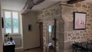 A room with a stone wall, a wooden ceiling, a dining area with chairs, a table, and a window with a view of greenery. - Appartement de Charme "Le Bureau des Armateurs" (Tréguier)