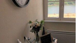 A room with a wall clock, a window with a view of a lake, a desk with a vase of flowers, and a small chalkboard. - Appartement de Charme "Le Bureau des Armateurs" (Tréguier)