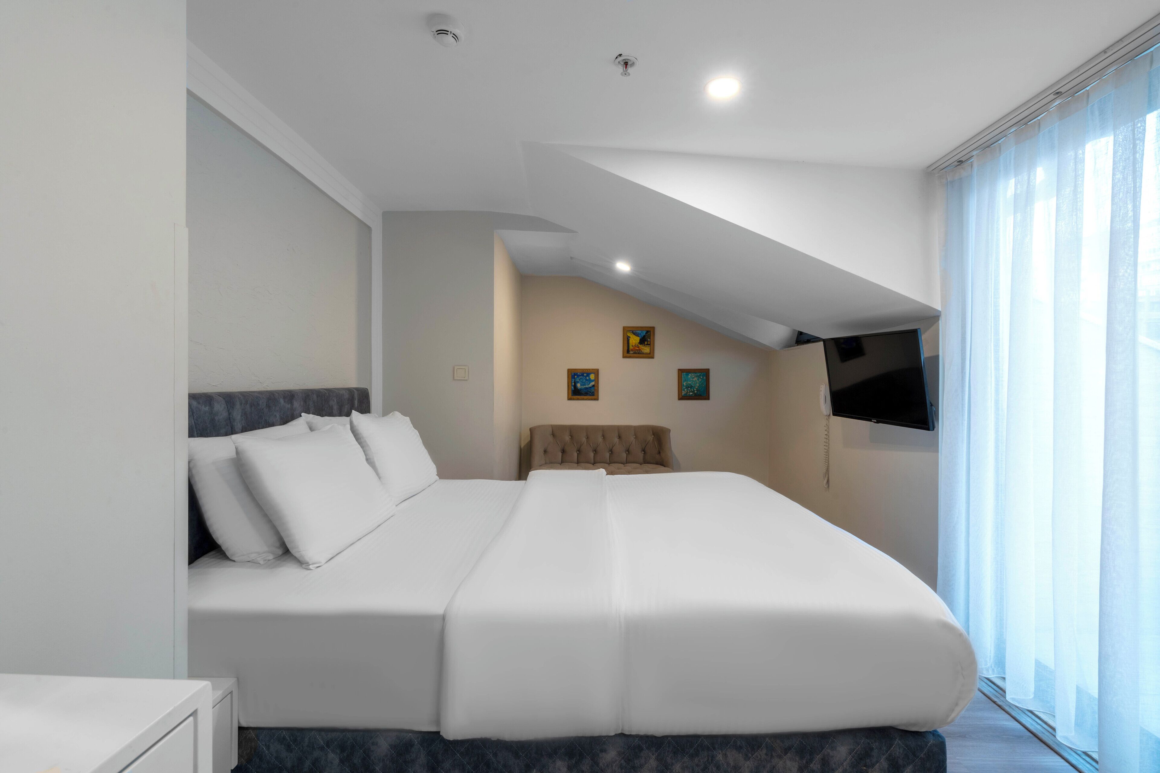 A modern bedroom with a large bed, a flat-screen TV mounted on the wall, and a window with sheer curtains.