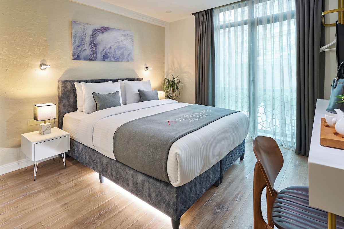 A modern hotel room with a large bed, a bedside table with a lamp, a desk with a chair, and a large window with curtains.