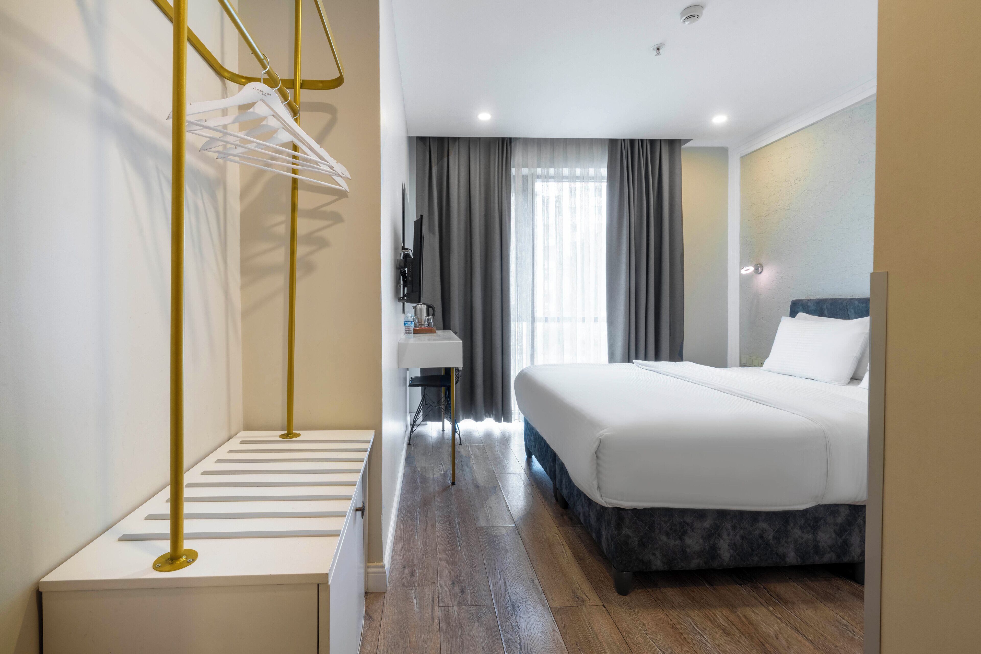 A hotel room with a bed, a desk with a chair, a hanging clothes rack, and a window with curtains.