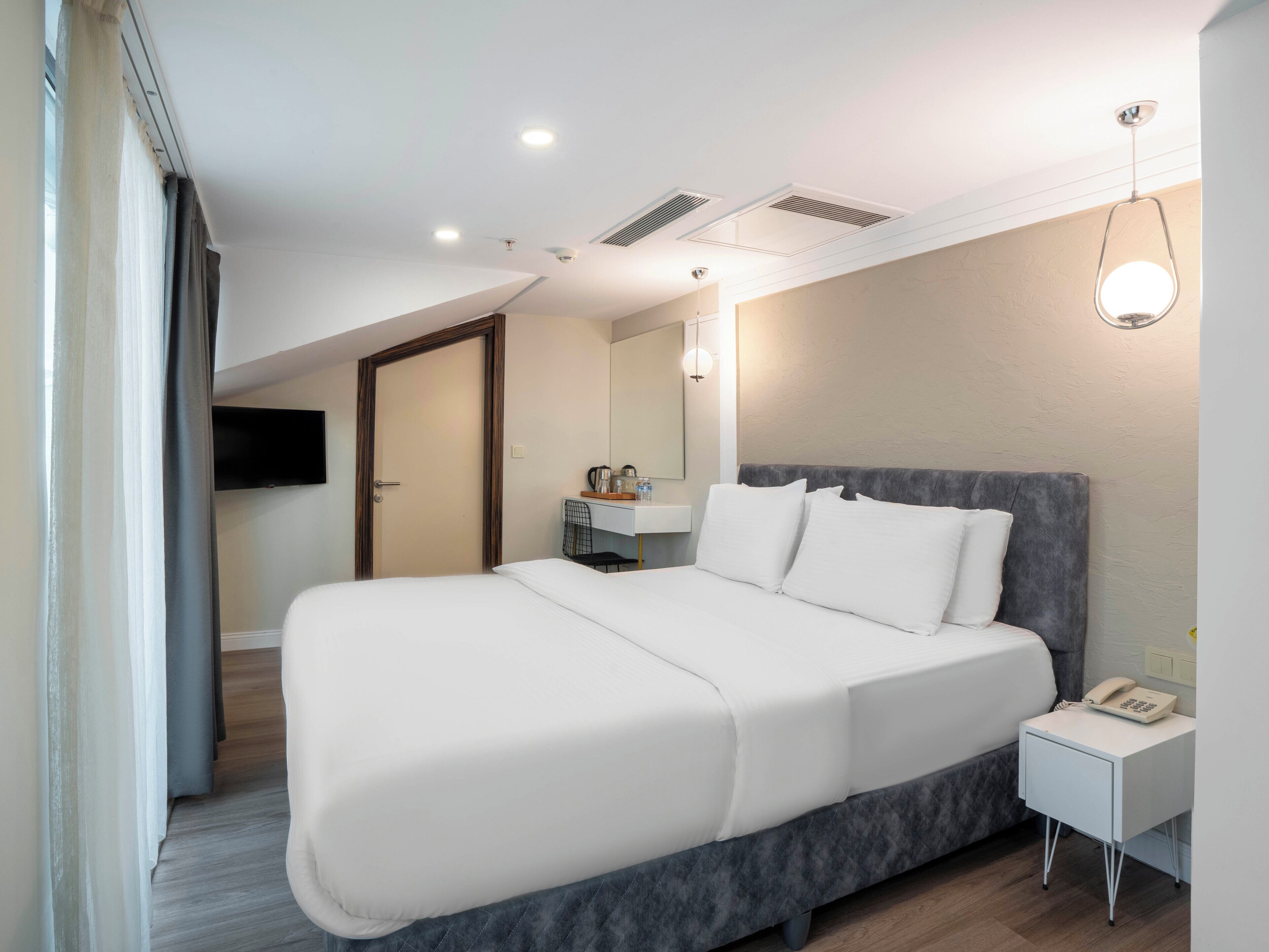 A modern hotel room with a large bed, a bedside table with a phone, a small desk with a mirror, and a ceiling light fixture.