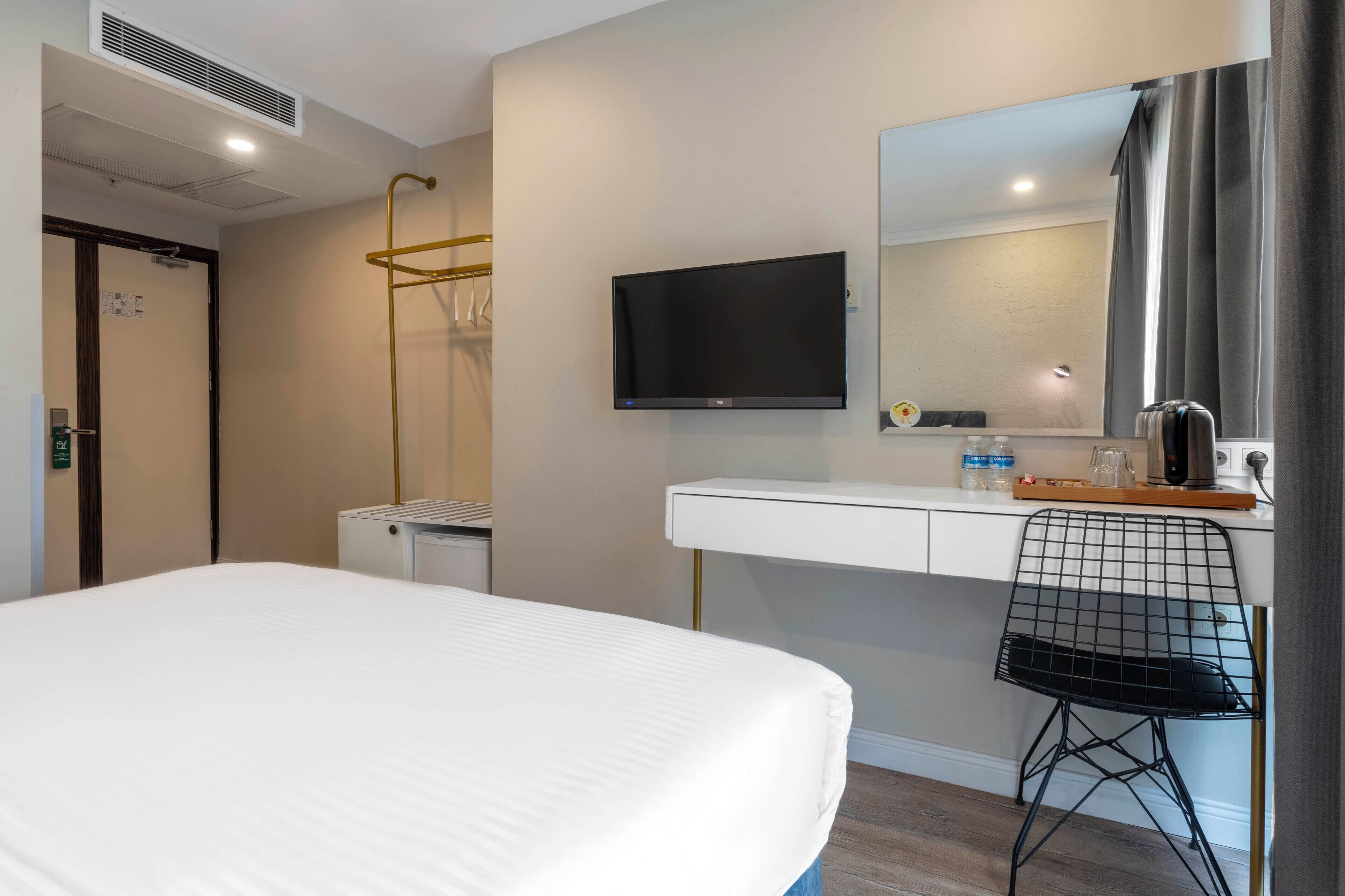 A hotel room with a bed, a desk with a chair, a TV, a mirror, and a window with curtains.