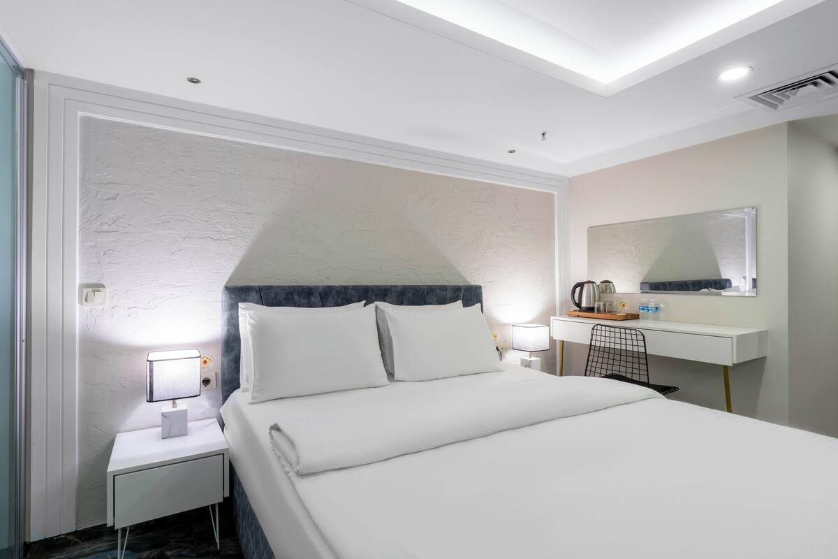 A hotel room with a neatly made bed, a bedside table with a lamp, a desk with a mirror, and a chair.