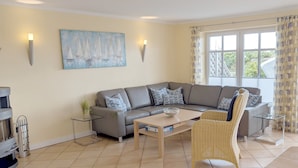 Un salon moderne avec un canapé d’angle gris, une table basse en verre, un fauteuil en osier et un tableau représentant des voiliers accroché au mur.
