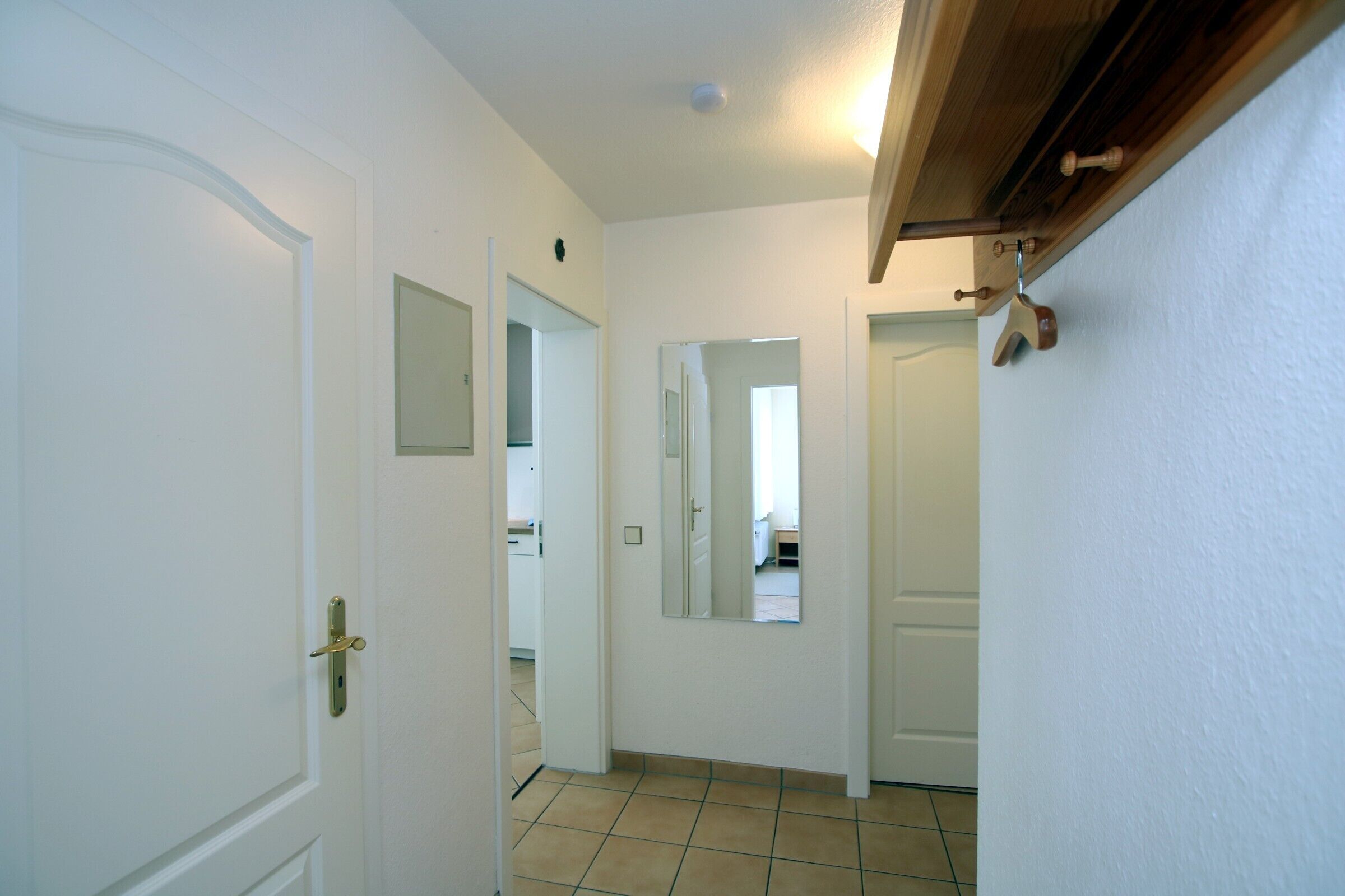 A hallway with white walls, a tiled floor, and a wooden ceiling. There are doors on both sides, and a mirror on the right wall.