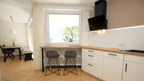 A modern kitchen with white cabinets, a wooden countertop, and a built-in microwave. There is a window with white curtains, a dining area with a table and chairs, and a radiator under the window.