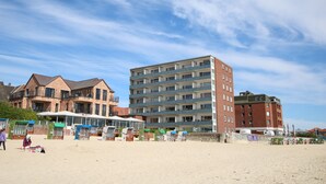 Una spiaggia con file di sedie a sdraio e ombrelloni, un grande edificio in mattoni con numerosi balconi, e un cielo sereno.