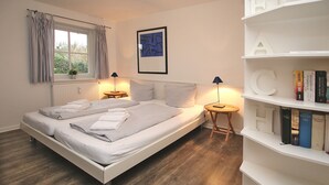 A neatly made bed with pillows and a bedside table with a lamp, a bookshelf with books, and a window with curtains.