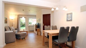 A modern living room with a sofa, ottoman, and dining table with chairs. There is a kitchen area with a TV and a window. - Ferienwohnung mit 3- Zimmern auf ca. 60m² im Erdgeschoß Eines Reihenhauses (Wyk auf Föhr)