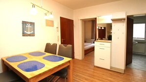 A room with a wooden dining table, chairs, a bed, a bathroom with a mirror, and a door leading to another room. - Ferienwohnung mit 3- Zimmern auf ca. 60m² im Erdgeschoß Eines Reihenhauses (Wyk auf Föhr)