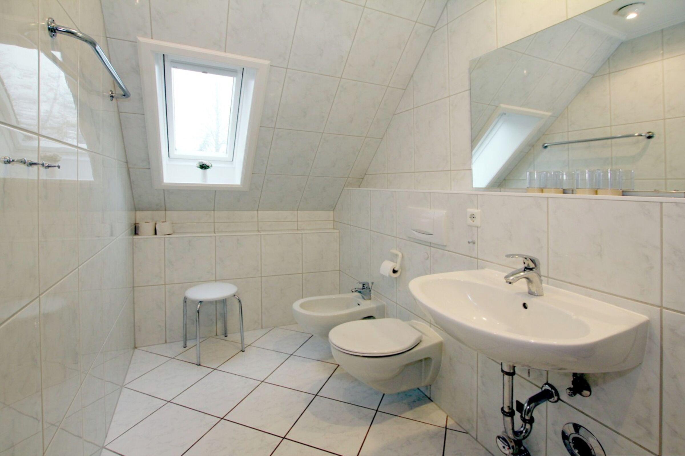 A modern bathroom with a toilet, bidet, and sink, tiled floor, and a window providing natural light.