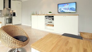 Une cuisine moderne avec des meubles blancs, un coin repas doté d’une table en bois et un téléviseur fixé au mur.