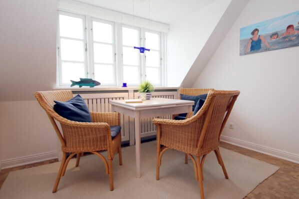 A room with two wicker chairs and a small white table, a window with a view of the sea, and a painting on the wall.