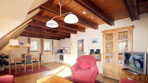 Ein gemütliches Wohnzimmer mit Holzboden, einem Essbereich mit Tisch und Stühlen, einem rosafarbenen Sessel, einem Fernseher und einer Vitrine.