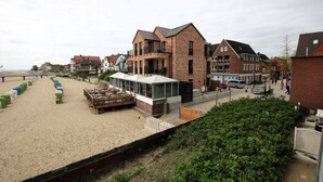 Un hôtel situé en bord de mer, doté d’une plage de sable, d’une terrasse en bois avec des sièges et de bâtiments traditionnels en briques.