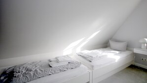 A small, white, single-bed room with a bedside table and a lamp.