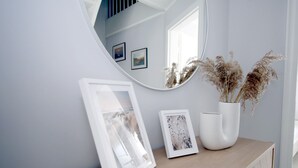 A wooden console table with a white vase of dried pampas grass, a framed photograph, and a mirror reflecting a staircase and framed pictures on the wall.