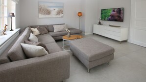 A modern living room with a grey sectional sofa, a matching ottoman, a small round coffee table, a white cabinet, and a flat-screen TV mounted on the wall.