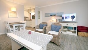 Una sala de estar acogedora con una mesa de comedor, dos sillones de mimbre, un televisor y una cocina al fondo.