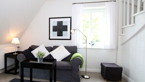 A modern living room with a grey sofa, a black coffee table, a small black ottoman, a floor lamp, and a window with white curtains. - Ferienwohnung mit 2- Zimmern auf ca. 35m² (Wyk auf Föhr)