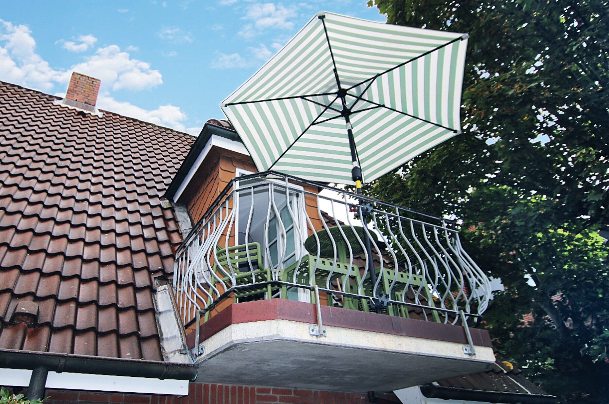 Ein Balkon mit einem grün-weiß gestreiften Sonnenschirm, einem Ziegeldach und einem Metallgeländer.