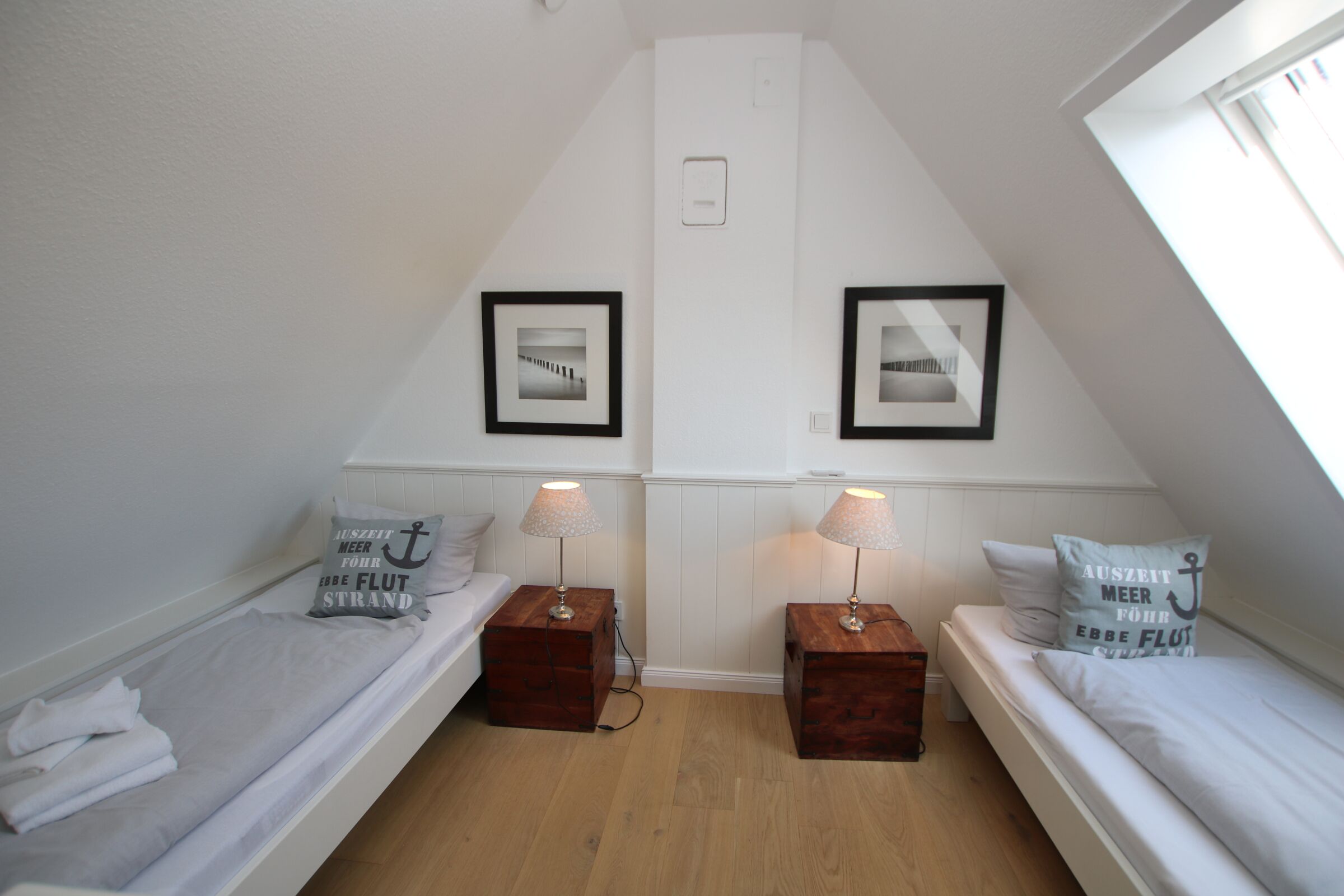Una habitación pequeña y bien iluminada con dos camas individuales, cada una con una mesita de noche de madera y una lámpara. Hay cuadros enmarcados en la pared sobre las camas, y la habitación tiene un techo inclinado.