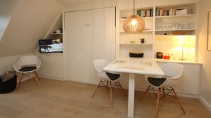 A modern dining area with a white table, two white chairs, a hanging light fixture, and a bookshelf with books and glassware. - Ferienwohnung mit 2- Zimmern auf ca. 35m² (Wyk auf Föhr)