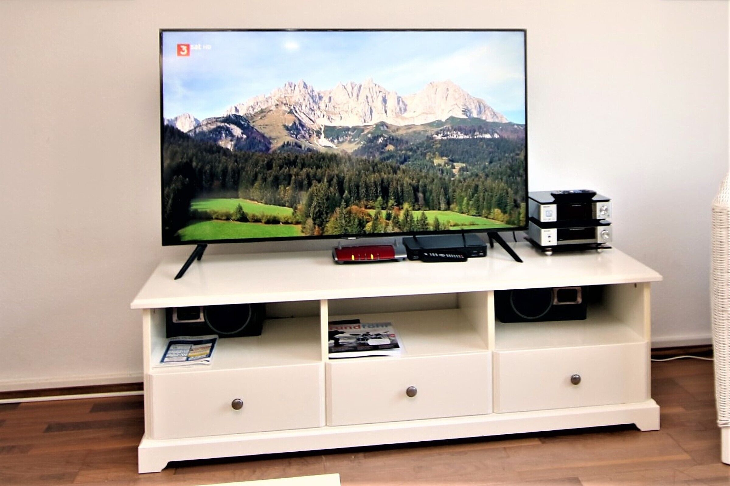 A flat-screen TV on a white entertainment unit with two speakers, a remote, and a magazine.