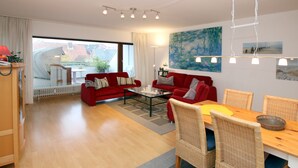 A modern living room with a red sofa, a dining table with wicker chairs, and a large window offering a view of the city.