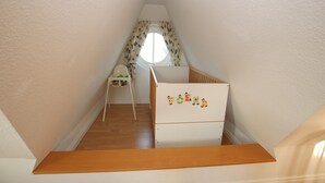 A small, narrow room with a wooden floor, a white crib with cartoon characters, a white chair, and a window with floral curtains. - Ferienwohnung mit 3- Zimmern auf ca. 55m² für Max. 2 Erwachsene und 2 Kinder (Wyk auf Föhr)