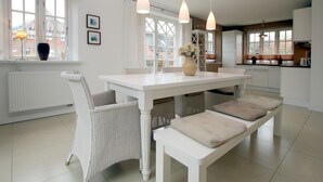 Una cocina luminosa con armarios blancos, una mesa de comedor blanca con bancos e iluminación colgante.