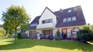 A two-story house with a large front lawn, a covered patio, and a garage. - Ferienwohnung am Südstrand von Wyk, mit 2 Zimmern auf ca. 44 m² im Erdgeschoß (Wyk auf Föhr)
