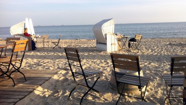 A beachside seating area with wooden and wicker chairs, a wooden table, and a white chair with a curved backrest. - Ferienwohnung am Südstrand von Wyk, mit 2 Zimmern auf ca. 44 m² im Erdgeschoß (Wyk auf Föhr)