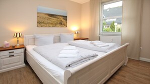 A neatly made double bed with white linens and towels, a bedside table with a lamp, a nightstand with a lamp, and a window with a view of greenery and a building. - Ferienwohnung am Südstrand von Wyk, mit 2 Zimmern auf ca. 44 m² im Erdgeschoß (Wyk auf Föhr)