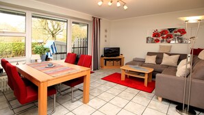 A modern living room with a dining table, red chairs, a sofa, a TV, and a large window. - Ferienwohnung in Wyk, mit 2- Zimmern auf ca. 42qm im Erdgeschoß (Wyk auf Föhr)