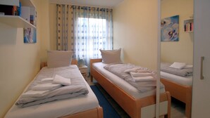 A hotel room with two single beds, a small bedside table with a lamp, a window with curtains, and a mirror reflecting the room. - Ferienwohnung in Wyk, mit 2- Zimmern auf ca. 42qm im Erdgeschoß (Wyk auf Föhr)