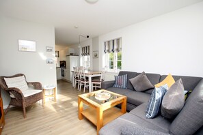 A modern living room with a grey sofa, wicker chair, and a wooden coffee table. A dining area with a white table and chairs is visible in the background.