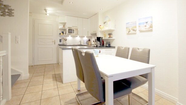 A modern kitchen with white cabinetry, a central island, and a dining area with a white table and chairs. - Reihenhausscheibe mit 3- Zimmern (Wrixum)