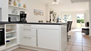 A modern kitchen with white cabinetry, a black countertop, and a coffee maker. There is a dining area with a chair and a table visible in the background. - Reihenhausscheibe mit 3- Zimmern (Wrixum)