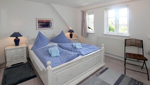 Un dormitorio con cama blanca, ropa de cama azul, mesita de noche con lámpara, una silla y ventana con cortinas.