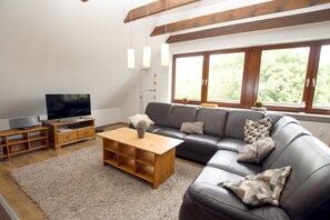 Ein Wohnzimmer mit einer Couch, einem Couchtisch, einer TV-Konsole und einem großen Fenster mit Blick auf Grünflächen.